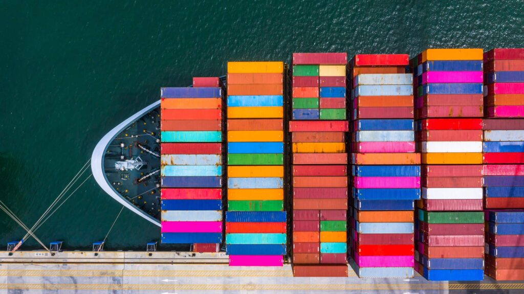 Aerial view of a large cargo ship docked at a port, loaded with colorful shipping containers neatly arranged in rows on its deck, as shipping documents are processed nearby over the greenish water.