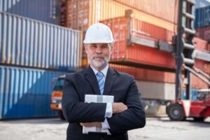 A man in a suit and white hard hat stands with arms crossed, holding a tablet displaying a final disbursement account, in front of stacked shipping containers and industrial machinery at a freight yard.