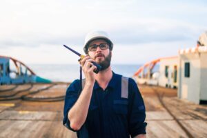 A man wearing a white hard hat, glasses, and a blue uniform speaks into a walkie-talkie on the deck of a ship, coordinating with port call templates as the ocean and equipment are visible in the background.