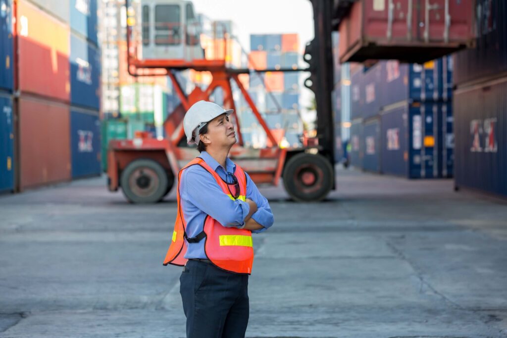 A person in a white hard hat, safety vest, and blue shirt stands with arms crossed, observing a shipping container being lifted—reflecting on the actual cost incurred in the busy yard filled with stacked containers.