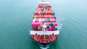 A large cargo ship loaded with colorful shipping containers sails through calm blue-green water, viewed from above and behind the bow, as its eta in shipping is tracked for timely arrival.