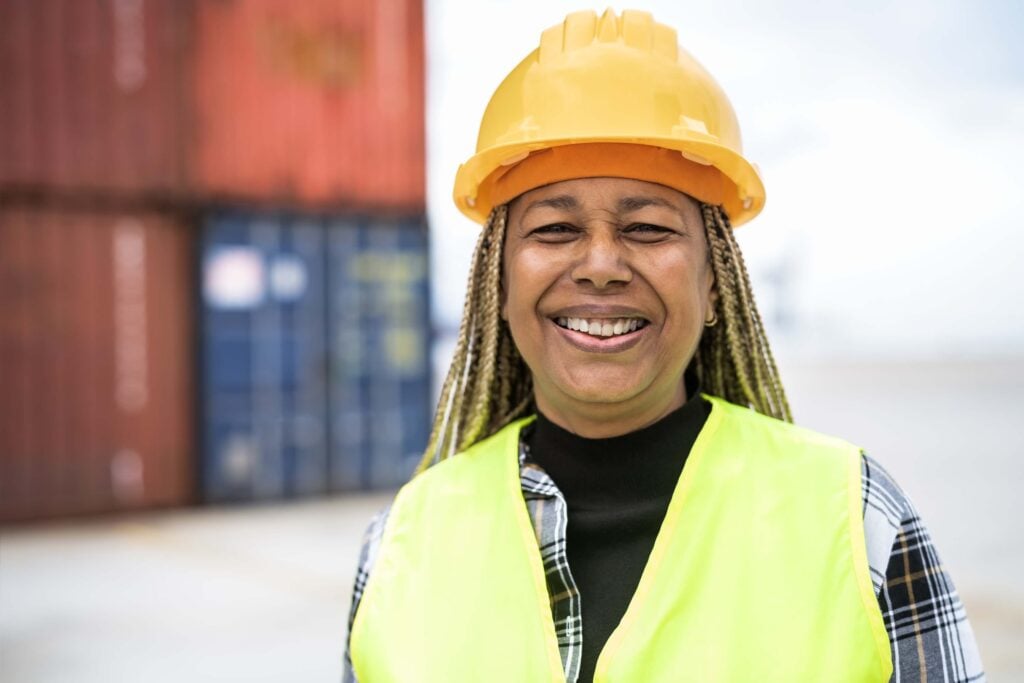 Smiling woman wearing a yellow safety helmet and neon vest stands outdoors in front of stacked shipping containers, highlighting her role in overseeing shipping companies and monitoring the estimated time (ETA) for cargo arrivals.