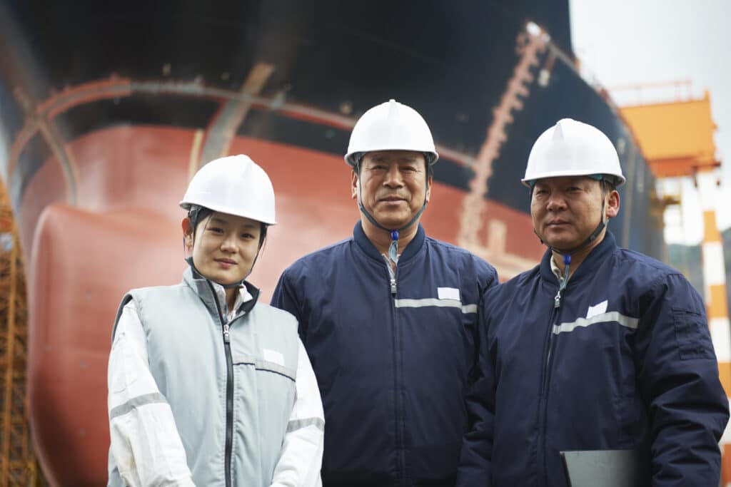 Three people in hard hats and work jackets stand before a large ship in a shipyard, where the total pilotage fee is discussed amidst the backdrop of the ship’s hull and industrial construction equipment.