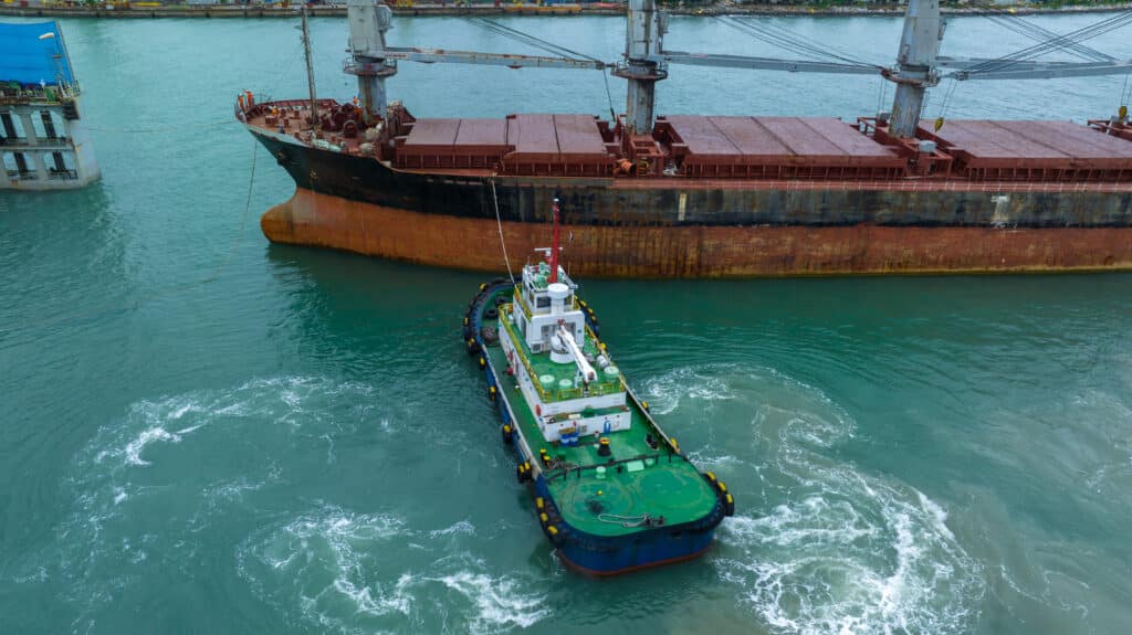 Aerial view of a small tugboat maneuvering near a large, rusted cargo ship in turquoise water at a port. The tugboat creates swirling patterns as it assists the vessel, with pilotage charge fees applying for this essential service.