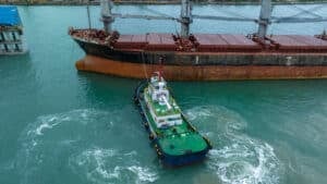 Aerial view of a small tugboat maneuvering near a large, rusted cargo ship in turquoise water at a port. The tugboat creates swirling patterns as it assists the vessel, with pilotage charge fees applying for this essential service.