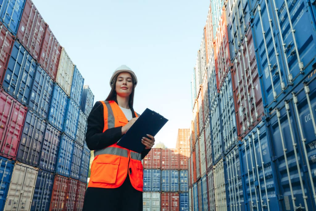 A woman wearing a hard hat and orange safety vest stands between stacks of shipping containers, holding port clearance documents on her clipboard as she looks ahead, likely inspecting or managing the cargo yard.