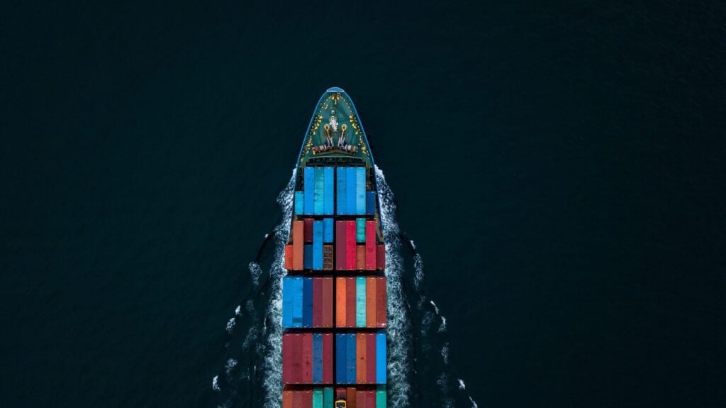 Aerial view of a large cargo ship sailing through dark blue water, carrying colorful shipping containers arranged in rows on its deck, en route to a busy port terminal for efficient cargo handling equipment operations.