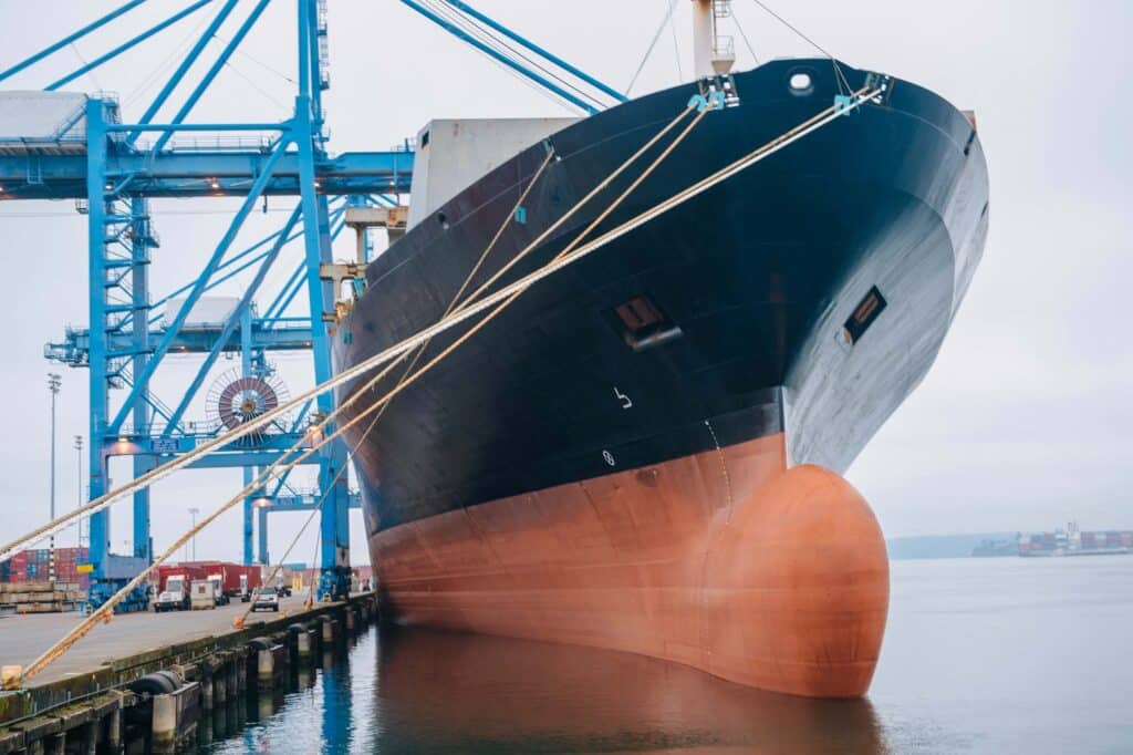 A large cargo ship docked at a port with thick ropes securing it. Blue cranes tower above, shipping containers are visible under an overcast sky, and crew members check communication equipment as part of the vessel agent checklist.