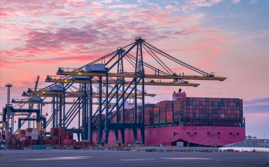 A large cargo ship loaded with colorful shipping containers is docked at a busy port terminal. Tall cranes and cargo handling equipment work efficiently at sunset, set against a stunning pink and purple sky in the background.