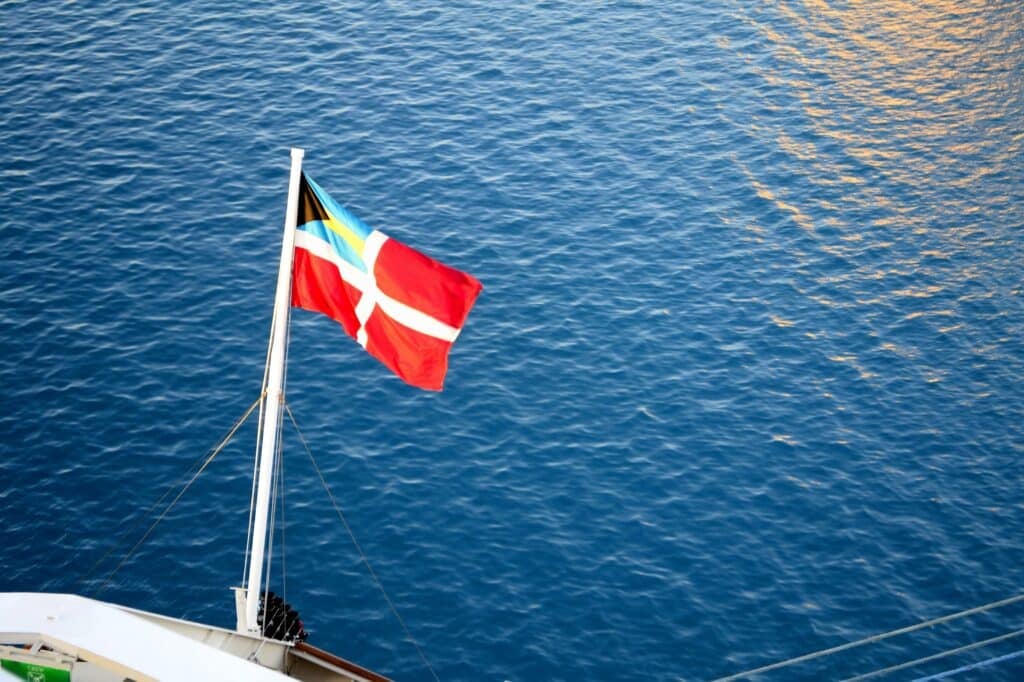 A red and white Scandinavian flag with a yellow and blue cross in the corner flies at the stern of a boat, symbolizing safety as enforced by flag state inspectors, with calm blue water in the background.