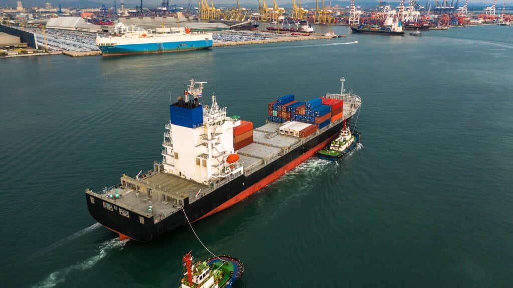 A large cargo ship carrying shipping containers is guided by tugboats in a busy port, showcasing efficient port operations with cranes, docked ships, and stacked containers visible in the background.