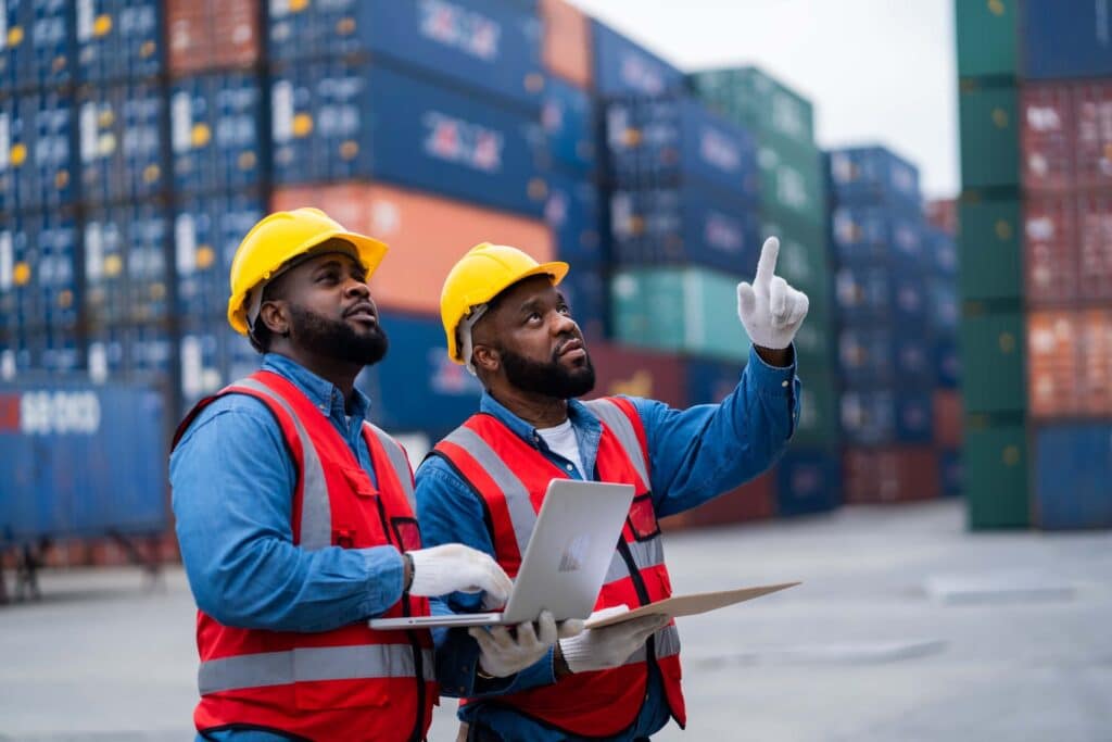 Two workers in safety vests and yellow helmets stand in a shipping yard with stacked cargo containers. One holds a laptop and clipboard while the other points upward, both engaged in port operations and area inspection.