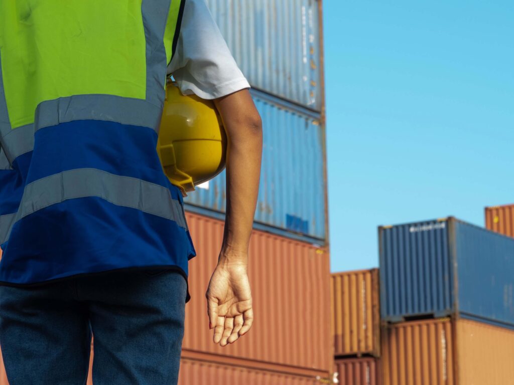 A worker wearing a safety vest holds a yellow hard hat while standing near stacked shipping containers, ready to assist with customized port calls under a clear blue sky.