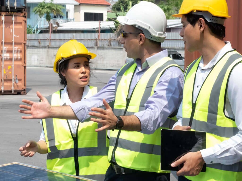 Three construction workers wearing safety vests and hard hats are having a discussion outdoors. On a worksite, one woman gestures as the two men, one with a tablet, listen—possibly coordinating with port service providers for a customized port call.