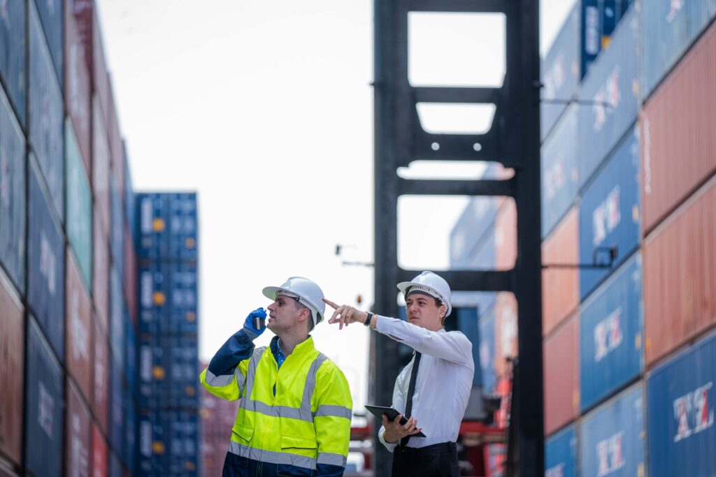 Two men in safety helmets and workwear stand among stacked shipping containers; one drinks from a cup while the other points ahead and holds a tablet, suggesting port call coordination at a busy shipping yard.