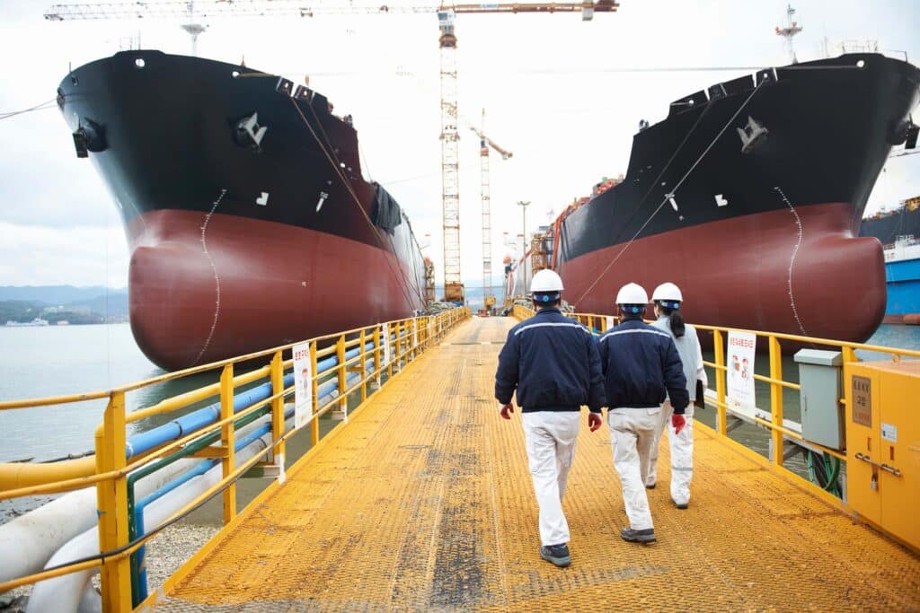 Three workers in uniforms and white helmets walk on a yellow dock between two large ships under construction, possibly reviewing a vessel agent checklist, with cranes and equipment visible in the background.