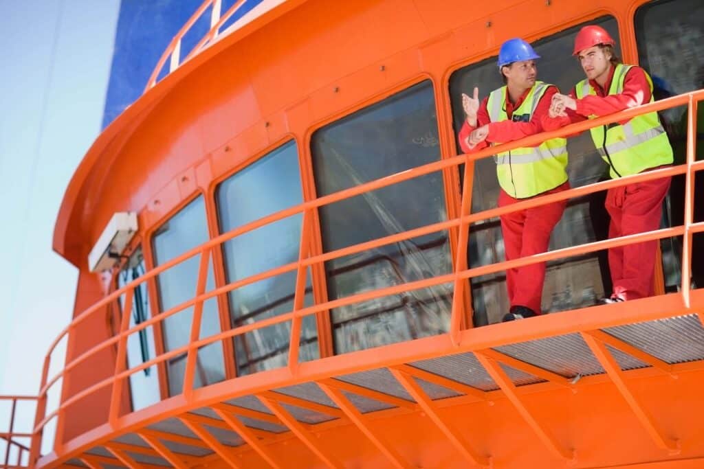 Two workers in red coveralls, yellow safety vests, and hard hats stand on the deck of an orange industrial structure, discussing communication equipment as they gesture, with large windows and a blue sky in the background.