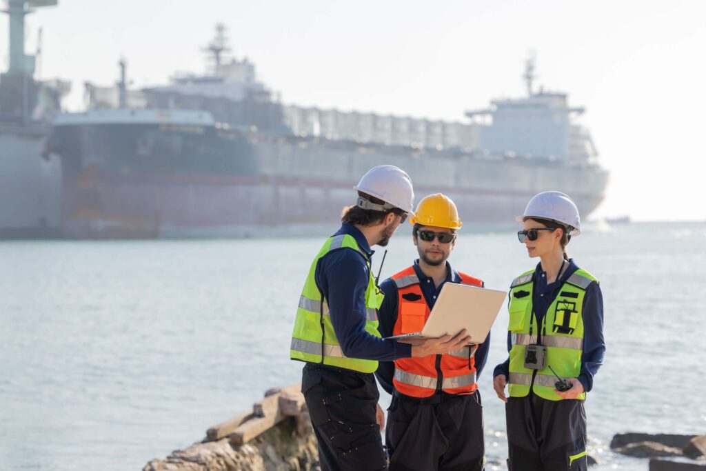 Three workers in safety vests and helmets stand near a waterfront, reviewing port operations on a laptop. A large cargo ship is anchored in the background under a bright sky.