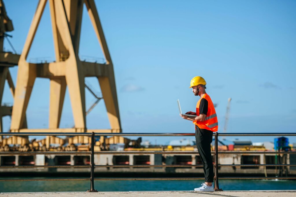 Dock worker using a laptop near a crane while monitoring port call changes and scheduling updates