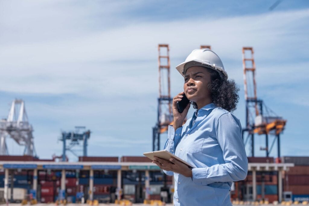 A vessel agent in a hard hat, phone in hand, holds a clip board at a port with cranes in the background.