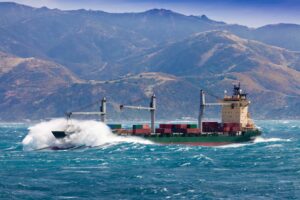 General average port call risk shown by a cargo ship pushing through heavy waves near a coastline