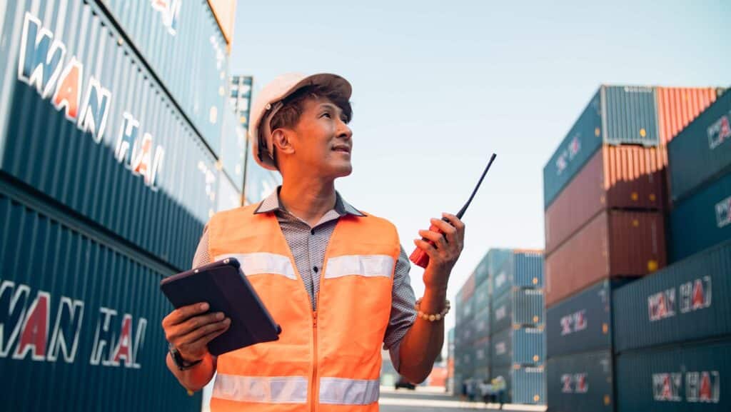Port agent vs ship agent duties shown by a port worker using a tablet and radio in a container yard
