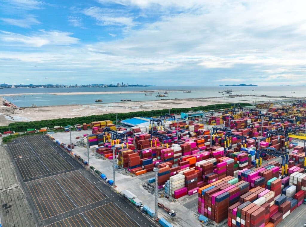 Aerial view of a busy container port with stacked cargo containers, cranes, trucks, and coastal water, representing port operations and maritime logistics.
