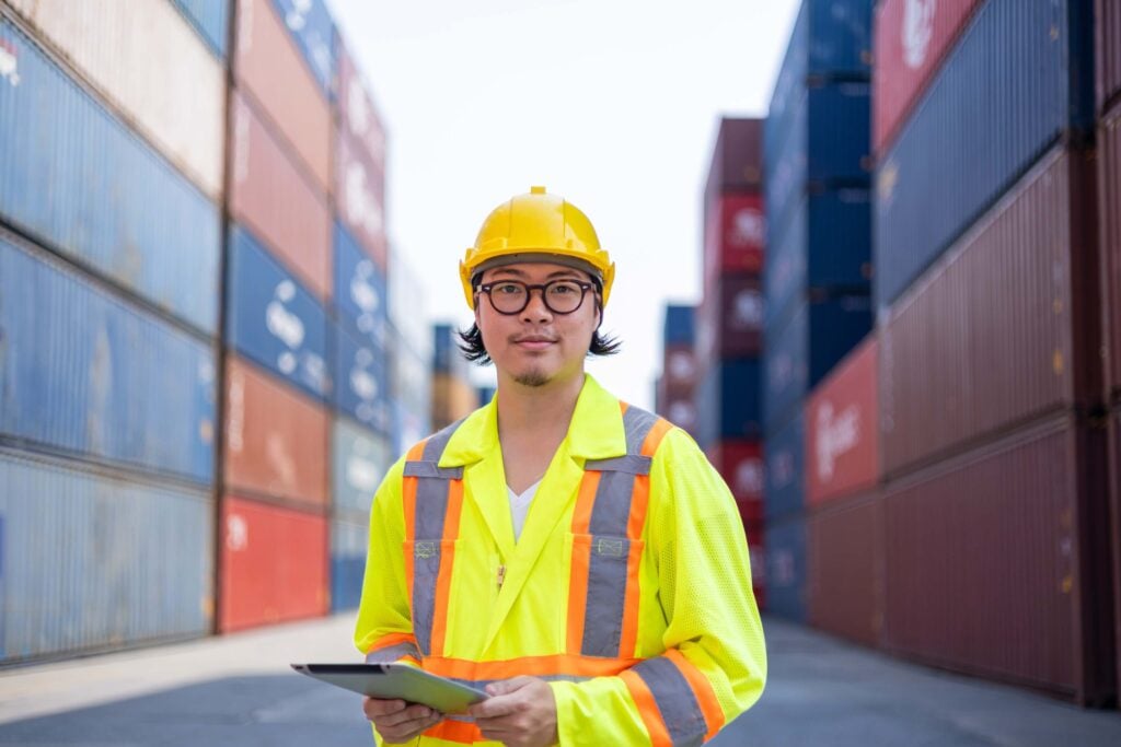 Port agent performance inspection by a logistics worker standing between stacked shipping containers
