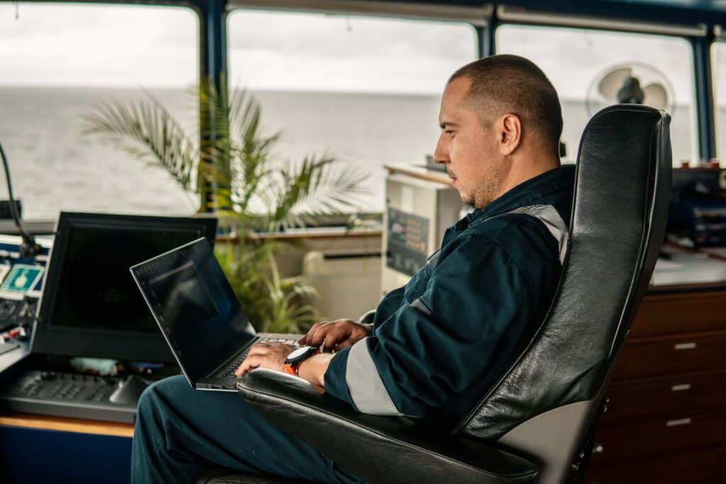 Navigational officer using a laptop on a vessel bridge while reviewing demurrage management software for live port-call visibility.