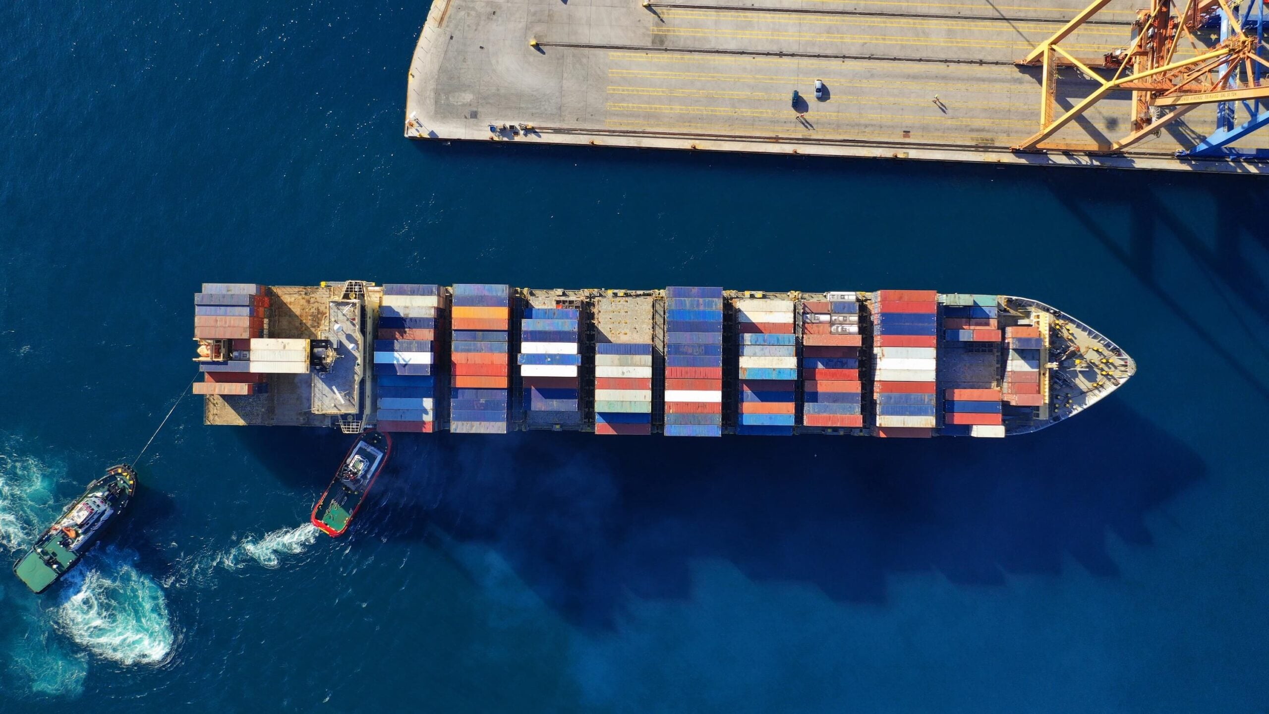 Aerial view of a large cargo ship loaded with colorful shipping containers, docked at a port with cranes, and a small tugboat guiding it through deep blue water—an important scene for vessel screening and maritime sanctions compliance in modern global trade.