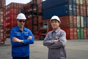 Two workers wearing hard hats and safety gear stand with arms crossed in front of stacked shipping containers at an industrial freight yard, highlighting the importance of maritime software solutions in streamlining logistics and ensuring safe, efficient operations.
