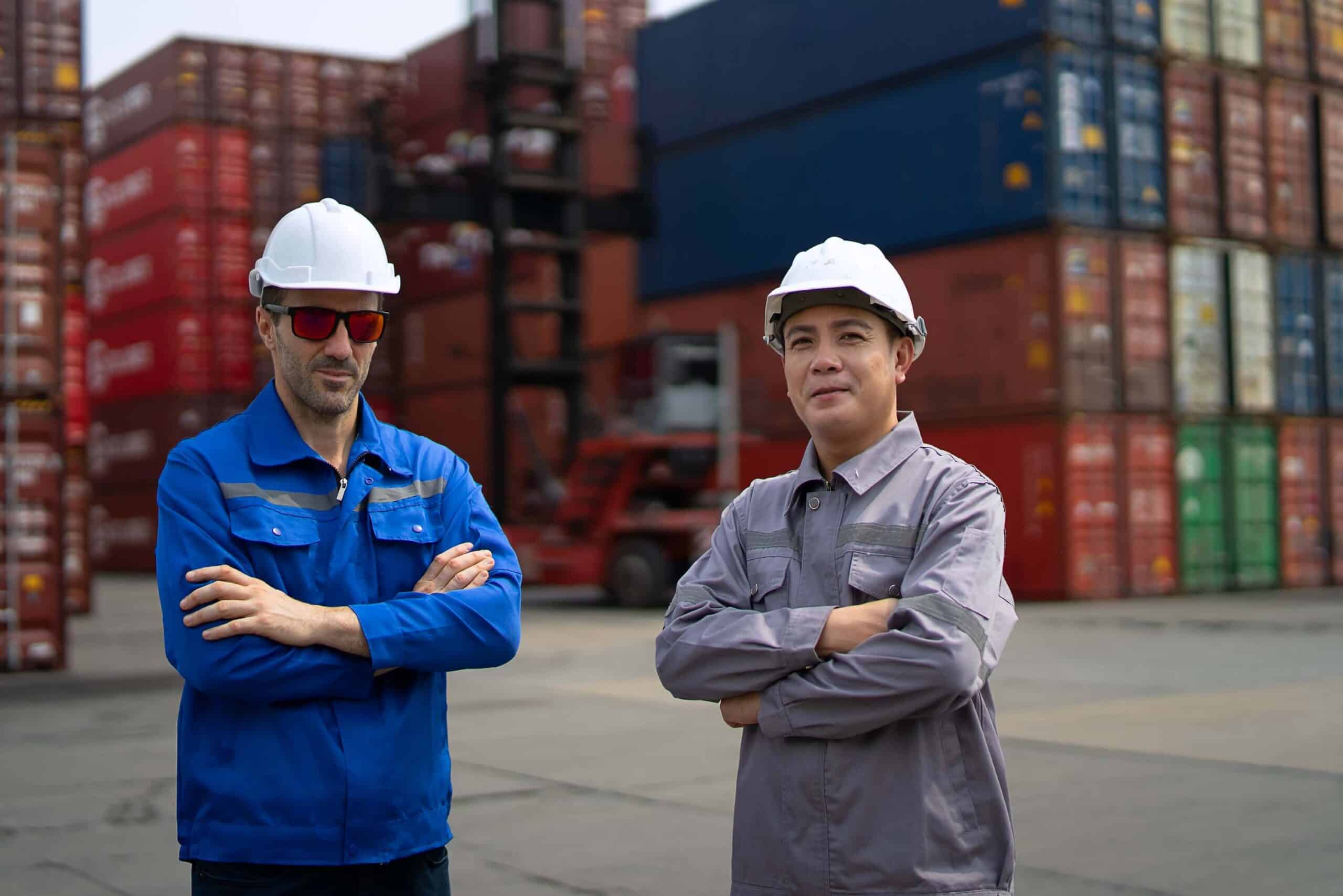 Two workers wearing hard hats and safety gear stand with arms crossed in front of stacked shipping containers at an industrial freight yard, highlighting the importance of maritime software solutions in streamlining logistics and ensuring safe, efficient operations.