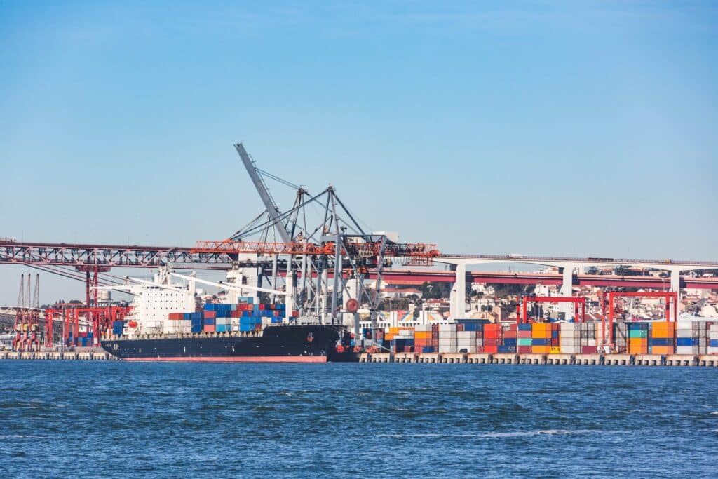 A cargo ship docked at a busy port with stacked colorful shipping containers, cranes, and a bridge in the background under a clear blue sky—a vibrant scene illustrating the efficiency and innovation brought by maritime software companies to the modern maritime industry.