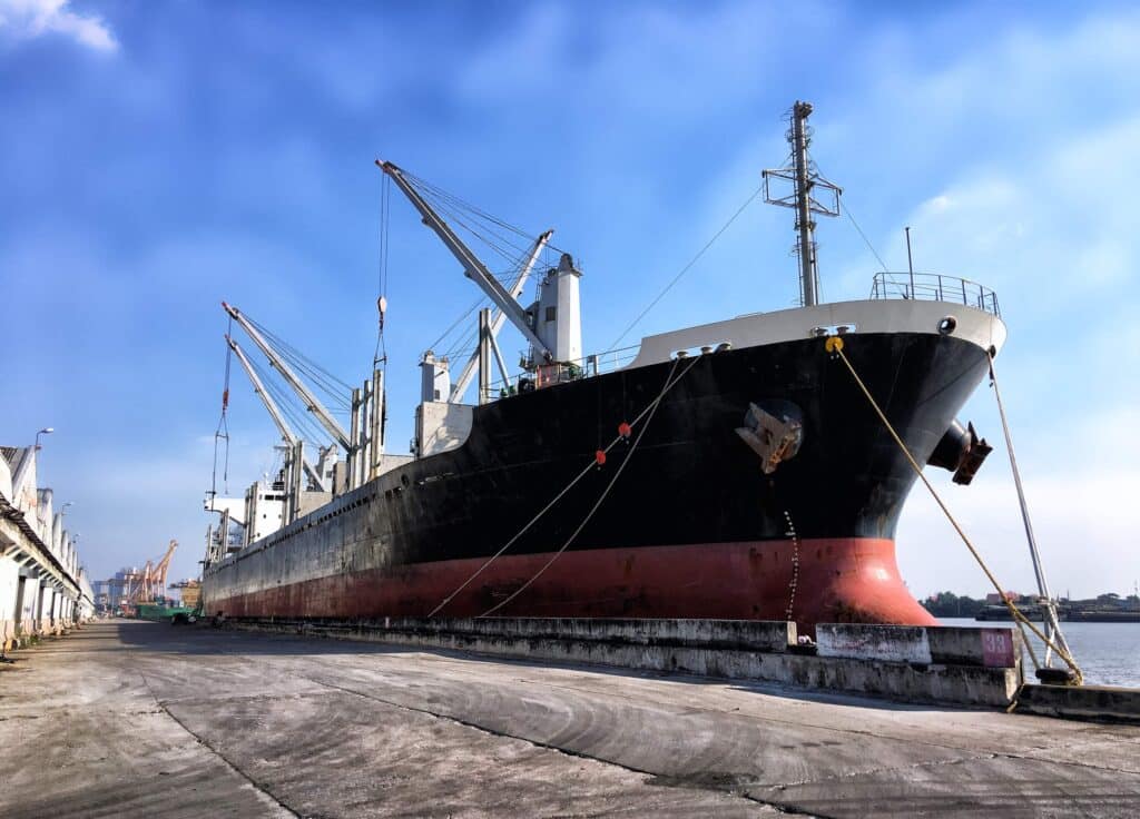 A large cargo ship is docked at a port, with cranes positioned around it for loading and unloading. The sky is clear and blue, and the dock area appears quiet and empty—a peaceful scene that highlights the efficiency possible with modern maritime software solutions.