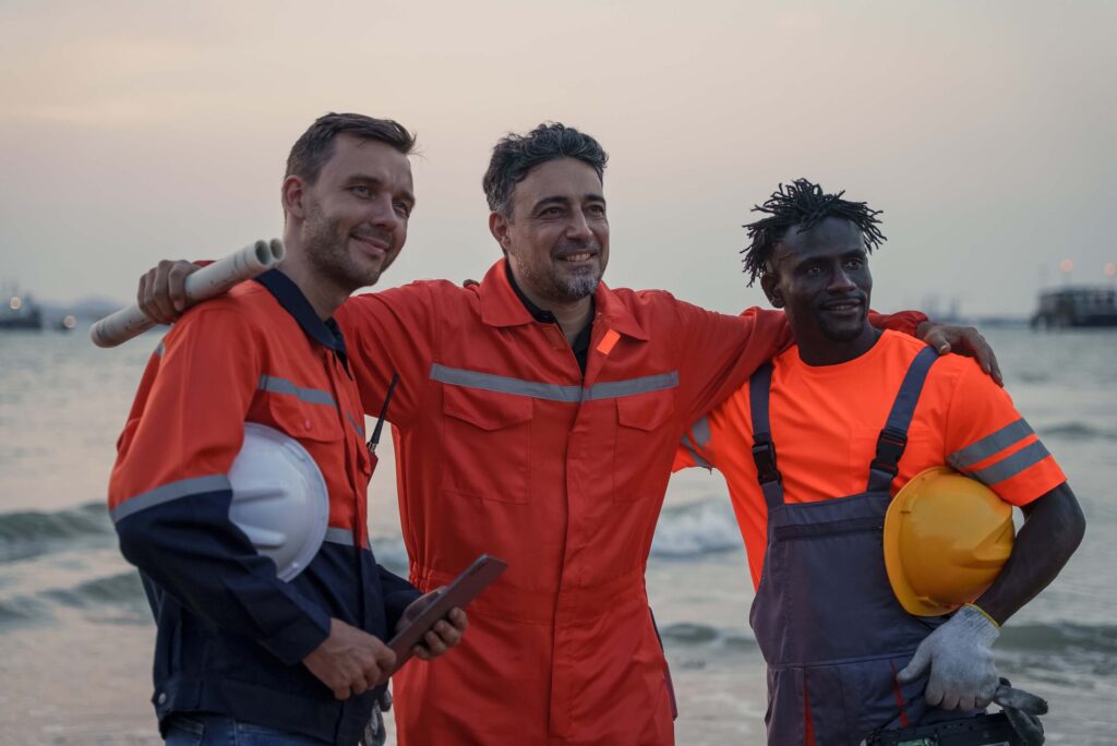 Three workers in orange safety gear stand together smiling on a beach at sunset, holding helmets and equipment, with the sea and sky in the background—embodying the teamwork essential for Marine Logistics Coordination for Offshore EPC Projects.