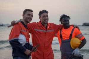 Three workers in orange safety gear stand together smiling on a beach at sunset, holding helmets and equipment, with the sea and sky in the background—embodying the teamwork essential for Marine Logistics Coordination for Offshore EPC Projects.