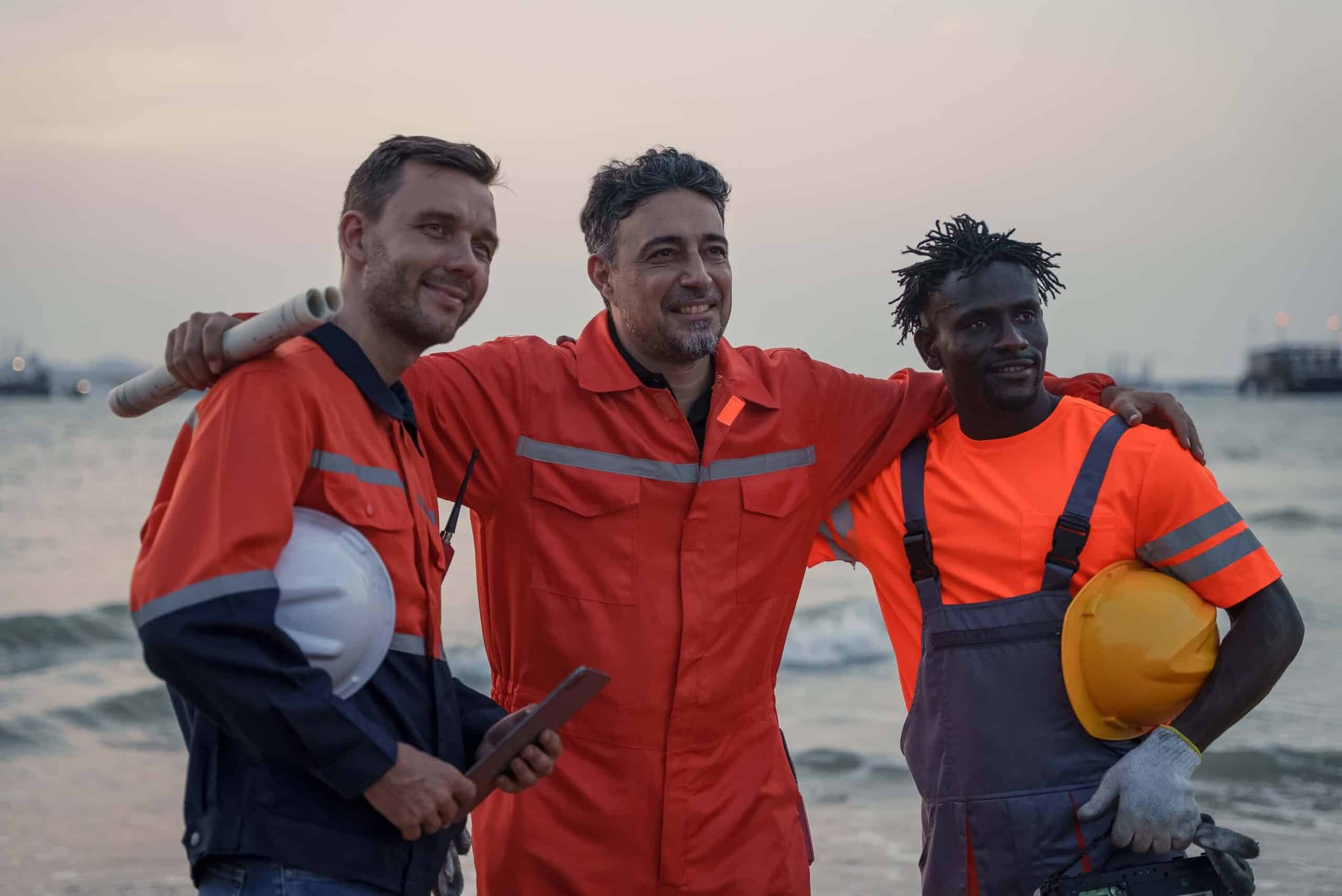 Three workers in orange safety gear stand together smiling on a beach at sunset, holding helmets and equipment, with the sea and sky in the background—embodying the teamwork essential for Marine Logistics Coordination for Offshore EPC Projects.