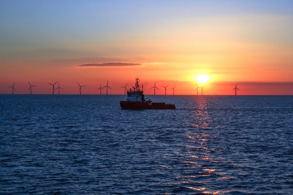 A boat sails on the ocean at sunset, with the sun low on the horizon. In the background, wind turbines—essential elements in offshore wind logistics—are visible, silhouetted against the sky. The water reflects the sunlight, creating a bright path towards the shore.
