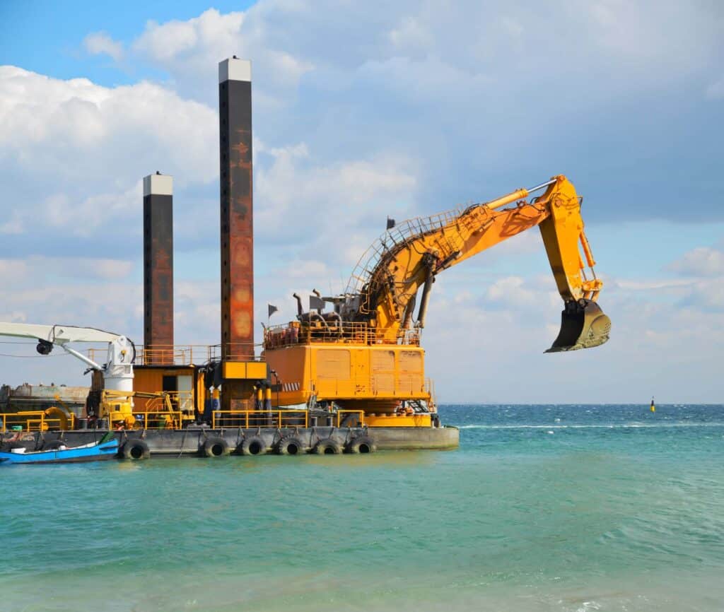 A large yellow floating excavator barge, positioned in turquoise water near the shore with its bucket arm extended, exemplifies effective marine logistics coordination. Two tall vertical supports rise from the platform under a partly cloudy sky, highlighting the vessel’s critical role in offshore EPC logistics operations.