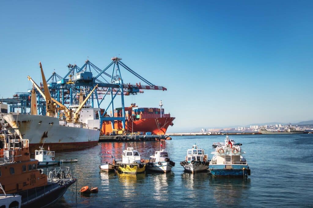 Several small boats float in a harbor near large cargo ships and blue cranes, illustrating the bustling activity typical of the maritime industry, with a cityscape and mountains visible in the background under a clear blue sky.