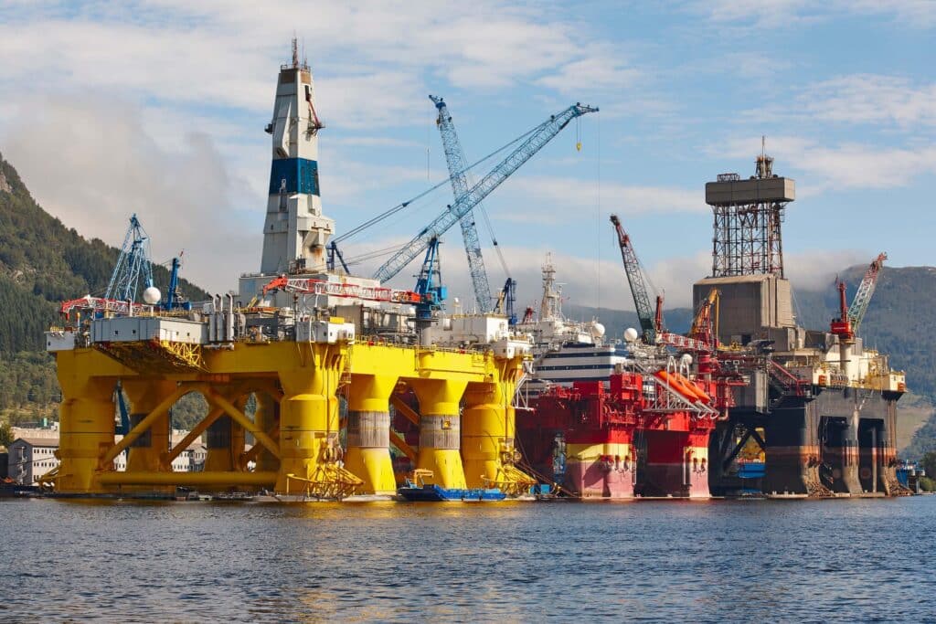Three large oil rigs, painted yellow, red, and black, are docked in a harbor with cranes surrounding them—an impressive scene of marine logistics coordination for offshore EPC projects. Hills and trees are visible in the background under a partly cloudy sky.