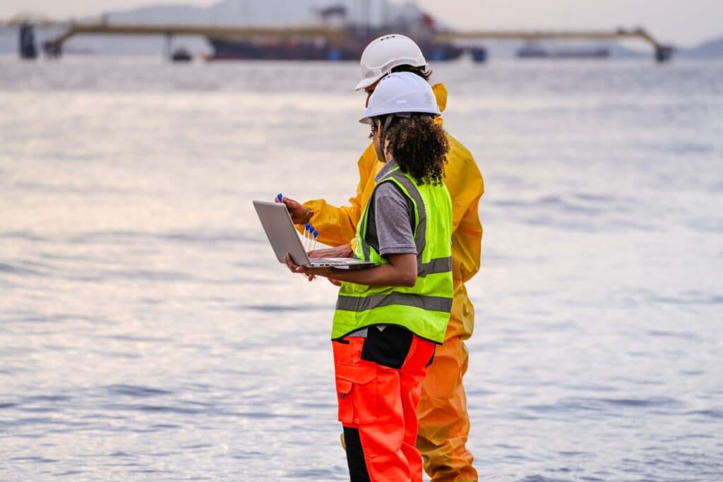 Two workers in safety gear stand by the water; one uses a laptop while the other gestures toward the sea, actively engaged in marine logistics coordination for offshore EPC projects. Both wear hard hats and high-visibility clothing. A ship is visible in the background.