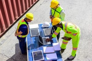 Three workers in safety gear examine solar panels and a laptop on a table outdoors, discussing data and plans. Clipboards and a hard hat are also on the table, as they review information relevant to maritime software companies focused on sustainable energy solutions.