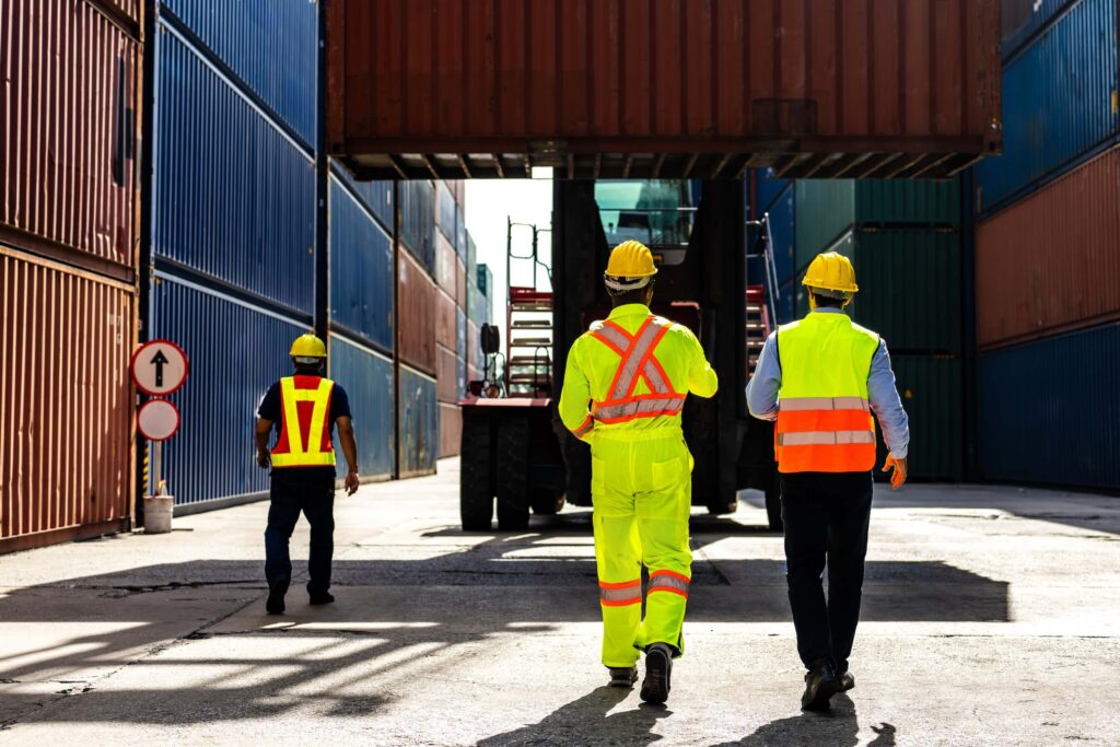 Three workers in safety vests and helmets walk through a shipping container yard, with a large container suspended above them by a forklift—a scene that highlights the importance of maritime software solutions in streamlining logistics and ensuring safety.