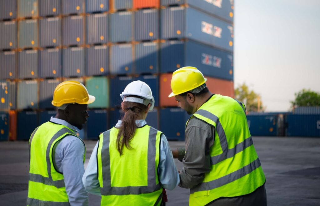 Three workers in safety vests and helmets stand together, talking in front of a stack of shipping containers at an outdoor industrial site, discussing the latest innovations from maritime software companies that are transforming the maritime industry.