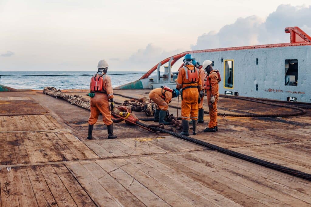 Four workers in orange safety gear and helmets handle equipment on the deck of a ship near large cables, demonstrating precise marine logistics coordination for offshore EPC projects, with the ocean and sky visible in the background.