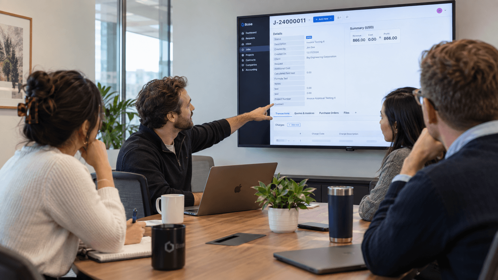 Four people sit around a conference table while one person points at a large screen displaying data and charts during a meeting in a modern office, offering a clear example of what to expect in a Base demo.