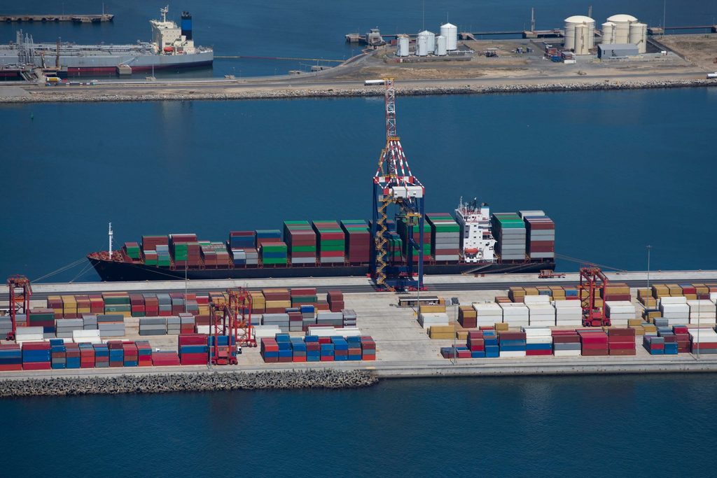 A cargo ship loaded with colorful shipping containers is docked at a port, with more containers stacked on the dock and cranes positioned nearby. In this bustling hub, Multi-Agent DA Reconciliation ensures efficient coordination among various operations. A body of water and storage tanks are visible in the background.