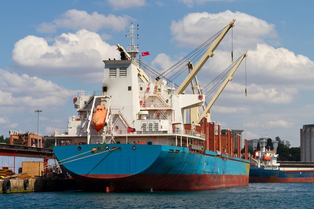 A large cargo ship docked at a port, with cranes on board and containers stacked on deck. Another ship and industrial buildings are visible in the background under a partly cloudy sky.