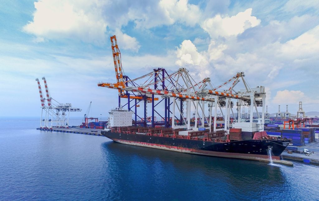 A large cargo ship docked at a busy port terminal with multiple cranes loading or unloading shipping containers under a partly cloudy sky.